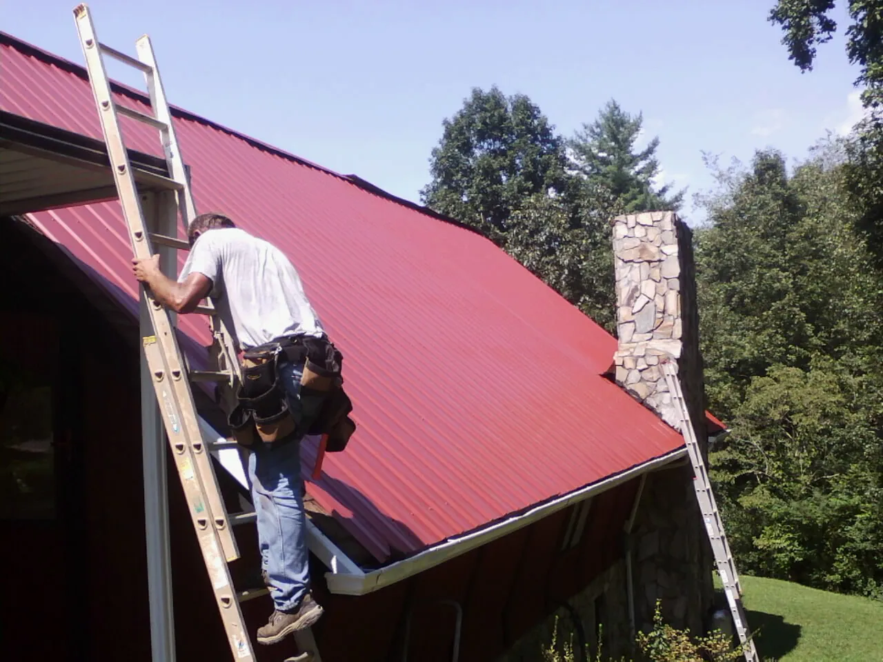 Roofer on a ladder beside a red metal roof and stone chimney — Soffit & Fascia Repair by Roofing Brothers in Sicklerville